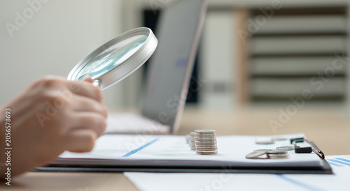 Tableau sur toile Business woman using a magnifying glass to examine financial reports and stacked coins on a desk, symbolizing audit, investment analysis, accounting, risk assessment, and business finance