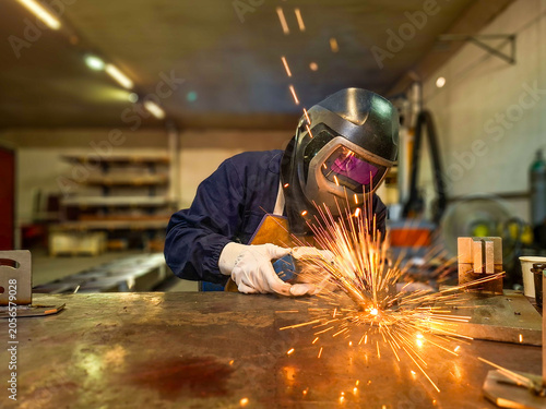 A professional welder wearing a protective mask and gloves works in a factory. Bright orange sparks fly out from the welding process on the metal table, contrasting with the blurred industrial worksho