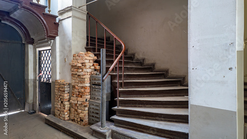 Obraz na plátně Neglected interior of a historic tenement house staircase with old wooden stairs, a metal handrail, a stack of bricks, mailboxes, and a decorative doorway portal on the left