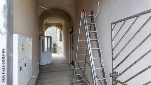 Interior of a renovated, bright tenement house corridor with arched ceilings, an aluminum stepladder leaning against the wall, white electrical boxes, and an open door leading to a courtyard.