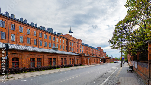 A long, historic red brick factory and former industrial complex Ksiezy Mlyn in Lodz, standing along an asphalt street with a sidewalk, trees, and a bench under a cloudy sky.