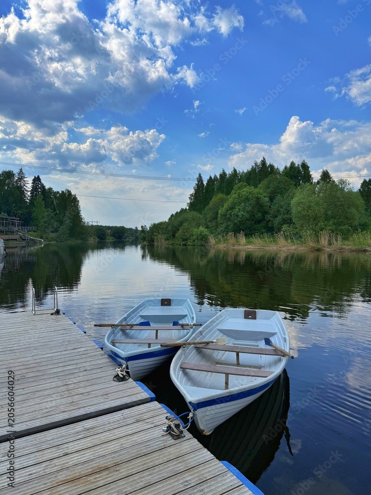 Obraz premium Two white rowboats with wooden oars are moored to a wooden pier on a calm river surrounded by forest under a cloudy blue sky. Concept of summer vacation and outdoor leisure.