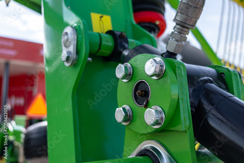 Macro view of a green-painted metal agricultural implement showing a bolted linkage, shiny hex bolts, and a hydraulic hose connection. Scene includes chipped