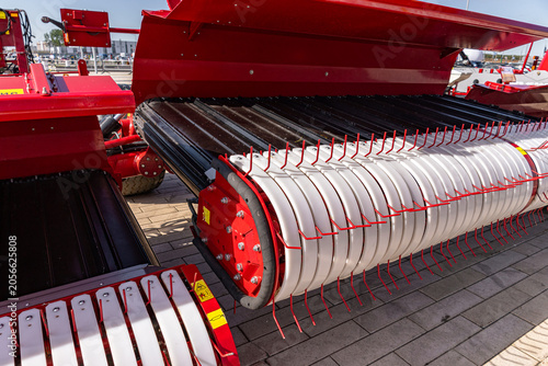 Close-up view of a red agricultural baler pickup assembly featuring metal tines, rotating roller drums, and painted steel housing. The scene shows interleaved