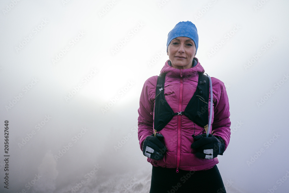 Fototapeta premium Confident female hiker in a pink jacket standing still on a misty mountain summit.Female Hiker Standing on Misty Peak