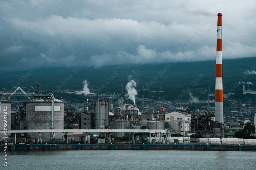 Fototapeta premium Industrial plant smokestack smoke factory refinery harbor storm cloud pollution chimney Industrial plant with smokestack and smoke near harbor under storm cloud over mountain