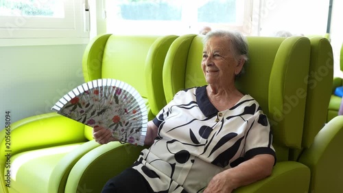 Smiling elderly woman sitting comfortably in a green armchair. Senior lady cooling herself with a hand fan inside a retirement facility