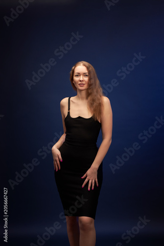 A woman with a beautiful figure in an elegant black dress poses against a dark blue background.
