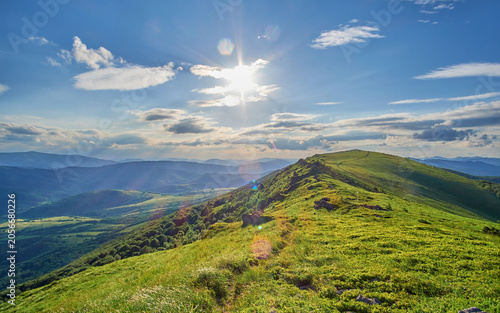 A dirt trail leads across a lush green mountain ridge toward a grassy peak, all under a vast, dramatic blue sky filled with bright, shifting white and grey cumulus clouds.