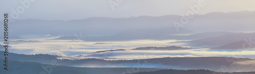 A panoramic view of rolling mountain ranges at dawn, with thick layers of ethereal morning mist blanketing the valleys and soft, hazy light bathing the distant peaks in a serene, peaceful atmosphere.