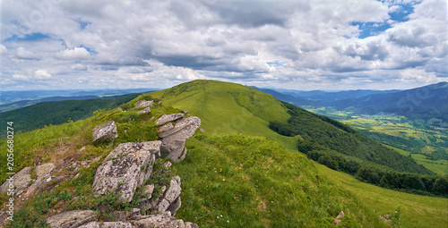 A dirt trail leads across a lush green mountain ridge toward a grassy peak, all under a vast, dramatic blue sky filled with bright, shifting white and grey cumulus clouds.