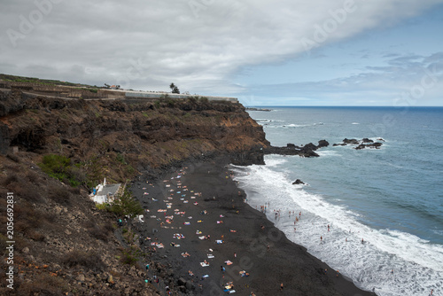 View of El Bollulo Beach, La Orotava, Tenerife, Spain, with black volcanic sand, gentle waves, and swimmers. 