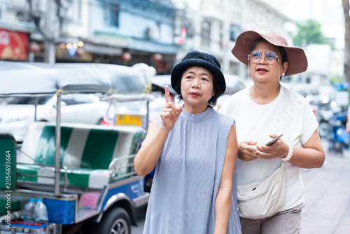 Senior asian women friends standing together on busy asian street beside tuk tuk looking around and talking about travel direction representing friendship adventure and retirement lifestyle