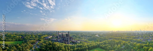 Eine beeindruckende Luftaufnahme des Landschaftsparks Duisburg-Nord zeigt die stillgelegten Hochöfen und Schornsteine des alten Hüttenwerks, umgeben von dichten, grünen Baumkronen.