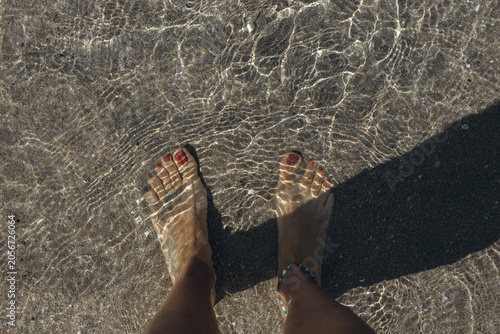 Top view of woman bare feet with red nail polish standing in crystal clear shallow sea water on a sandy beach, summer vacation POV in Sicily, Italy