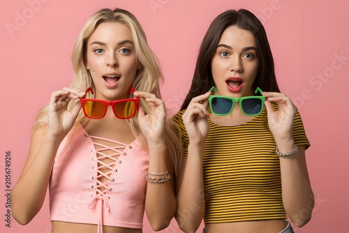 Surprised young women holding colorful sunglasses, stylish female friends with expressive faces, trendy summer fashion concept on pink background