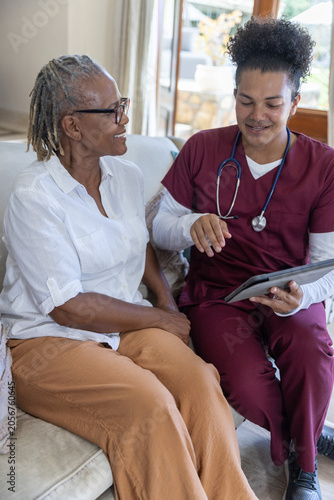 Nurse in scrubs showing tablet and stethoscope to Senior African American woman on couch at home