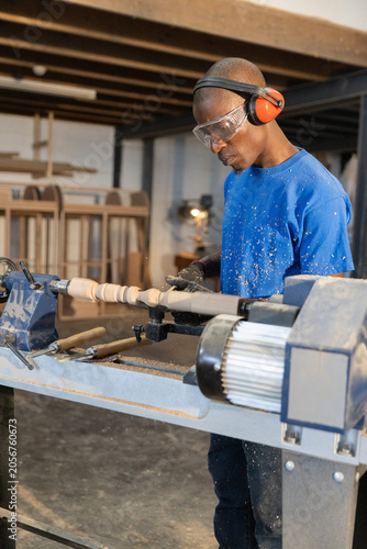 Woodworker shaping wood spindle on lathe in workshop with chisels, wearing goggles and headset