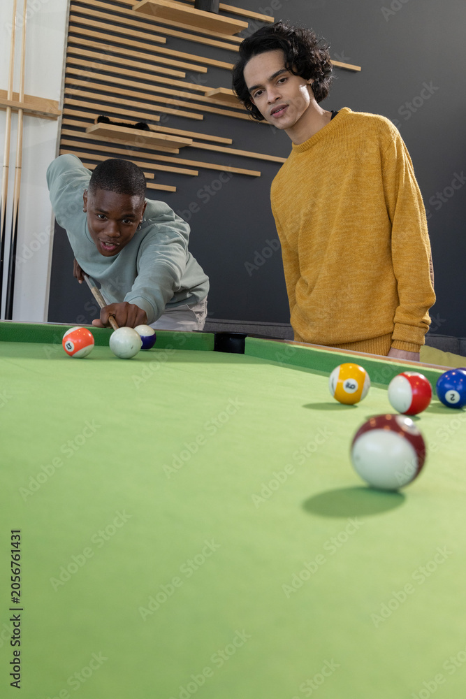 Obraz premium African American and Asian men playing pool in lounge on green-felt table, aiming among pool balls