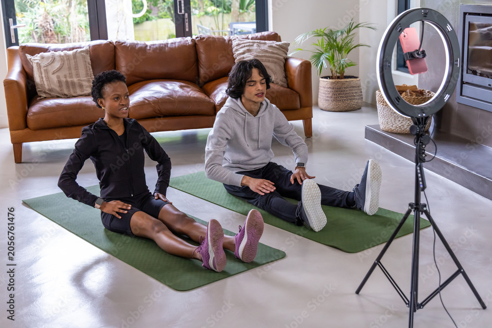 Obraz premium Diverse duo in activewear stretching on green mats in living room, recording with ring light phone