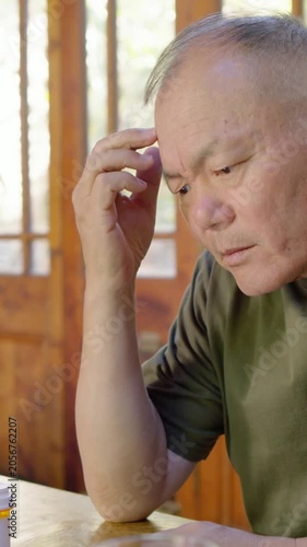 Vertical video: Seeing pill, senior in olive tee tapping table, rubbing temple and thinking at home