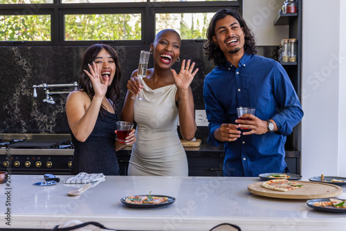 Diverse friends waving and holding drinks behind kitchen island, showing pizza slices and flute