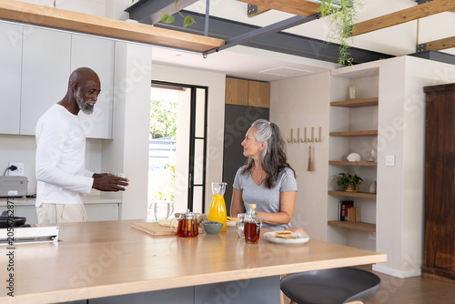 Diverse senior couple chatting at kitchen island with glass pitcher of orange juice and pastries