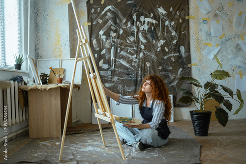curly woman painter creating masterpiece picture sitting on floor creative workshop studio near easel