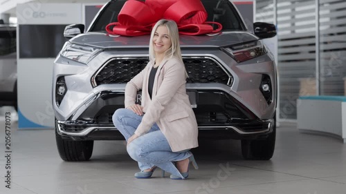 Happy business woman smiling and crouching next to new car with red bow in dealership