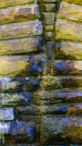 Textured stone wall of an old arched bridge featuring a dark V-shaped drainage channel through which water flows downward. Historic Byrd Creek Dam