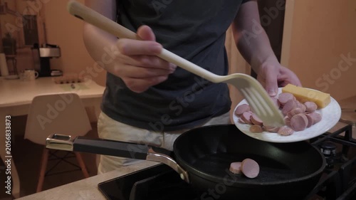 Hands adding sausage slices to skillet, casual home cook using spatula to flip browning pieces over hot oil, cozy apartment kitchen evening, quick breakfast or snack prep, savory sizzle and aroma