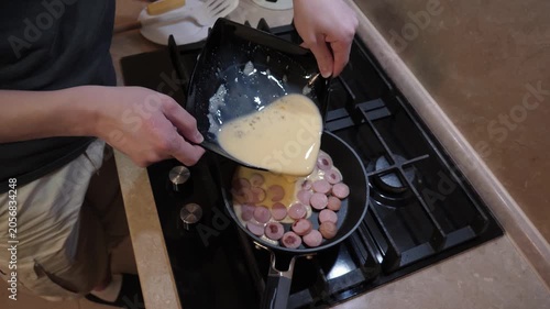 Homely egg and sausage preparation. Casual chef prepares eggs and sausage in cozy apartment kitchen. Weekend cook expertly adding eggs into frying pan alongside sliced sausages