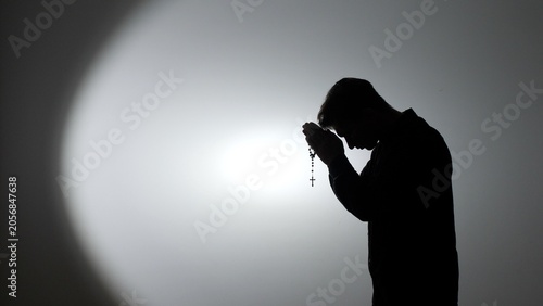 Obraz na płótnie Man's silhouette bending in prayer, holding rosary beads with a cross, highlight