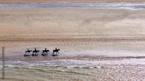 Horseback riding on wide sandy beach in Deauville, Normandy, France. Riders moving near surf line in aerial seaside view