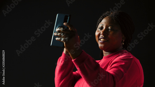 Tableau sur toile A cheerful woman in black background waving at a video call on her smartphone having a faraway interaction on an internet videoconference, with discussion on mobile phone via video conferencing