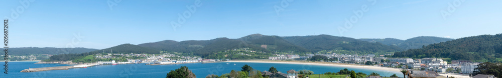 Fototapeta premium A sweeping panoramic view of Viveiro town curved around the sandy bay and blue sea of the Covas beach with rolling green mountains under a clear sky in Galicia, Spain.