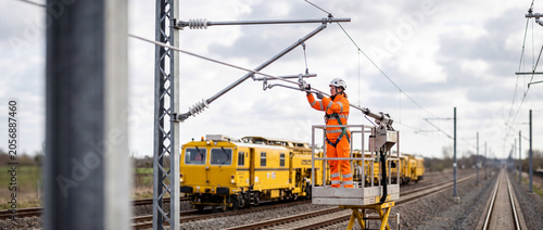 Railway worker performing overhead line maintenance on electrified tracks beside yellow service train