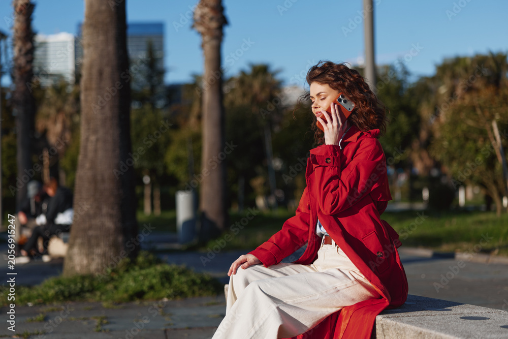 Obraz premium Young woman with curly hair wearing a red jacket talks on a smartphone while sitting on a bench in a sunny urban park with palm trees in the background.