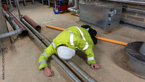 Construction engineer in high visibility coat and hard hat has tripped over a pipe in a plant room.  Slips, trips, and falls at work affect health and safety in the workplace