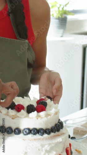 Vertical video: Squeezing piping bag baker in green apron finishing 2tier cake with berries at home