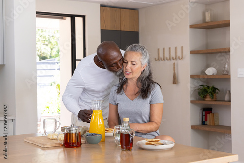 Diverse senior couple leaning over kitchen island, sharing breakfast with orange juice pitcher