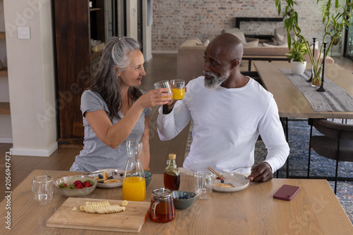 Diverse mature couple raising clear glasses of orange juice at home dining table with glass pitcher
