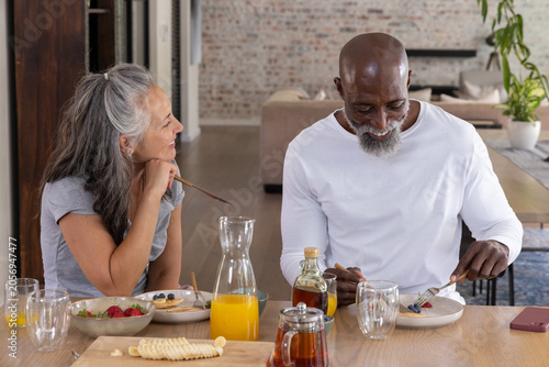 Senior woman leaning on hand and man cutting pancakes at wooden dining table with orange juice