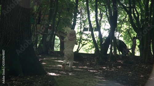 Shot from behind, a baby girl in a pink dress and a sunhat holds a large long stick walks along path forest among trees, background she sees mother tree house and begins run amusingly