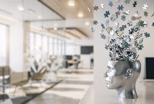 Silver metallic human head profile sculpture erupting with shiny floating jigsaw puzzle pieces. Blurred modern office background provides functional copy space.