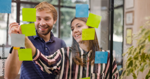 Two individuals work together on a project, placing and moving sticky notes on a glass wall in a bright office. They appear engaged as they share ideas and plan