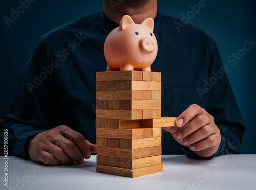 Piggy bank balancing on wooden block tower while a person removes pieces in a risky game concept. Financial stability, investment risk, savings protection and economic uncertainty in personal finance.