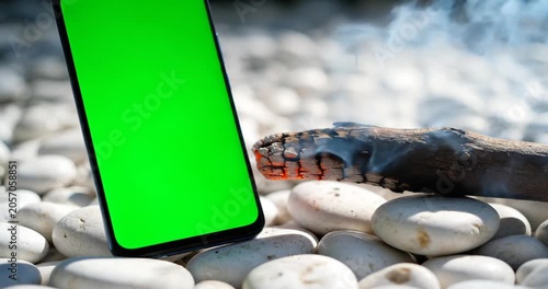 Smartphone with green screen placed on smooth pebbles near a glowing log in nature