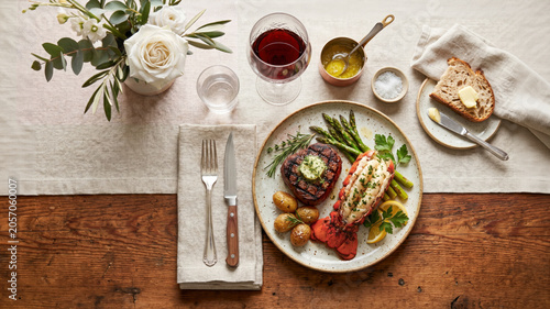 A flat lay of an elegant dinner featuring grilled steak, lobster, asparagus, and red wine on a wooden table. Elegant surf and turf setting.
