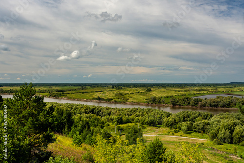 A river flows through a grassy green plain, the sky is covered with clouds, which gives the scene a dramatic atmosphere.